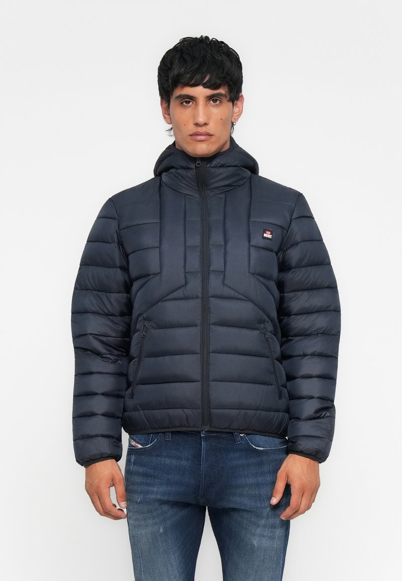 Junger Mann in einer schwarzen Steppjacke mit Kapuze und blauer Jeans, der vor einem schlichten weißen Hintergrund steht.