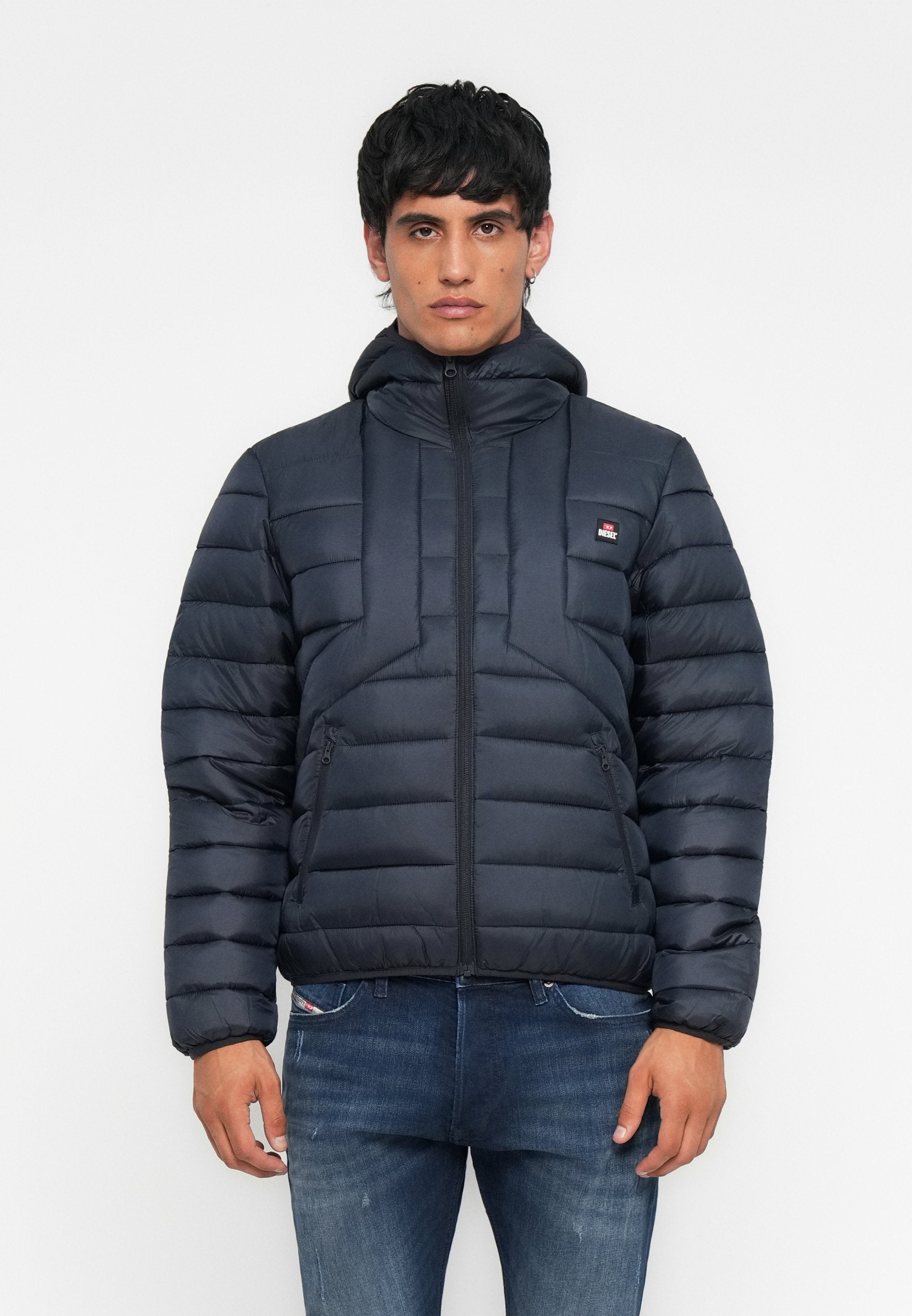 Junger Mann in einer schwarzen Steppjacke mit Kapuze und blauer Jeans, der vor einem schlichten weißen Hintergrund steht.