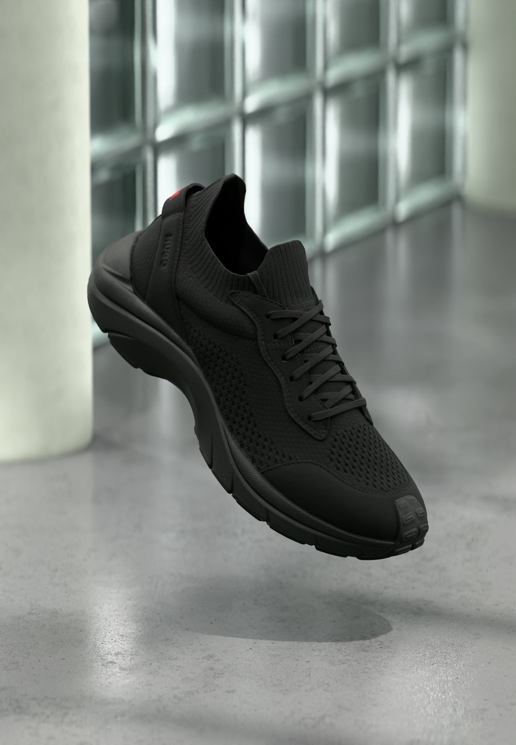 Schwarzer Sportschuh mit gestricktem Obermaterial, Schnürdesign und gepolsterter Sohle, der über einem polierten Betonboden in der Nähe von Glasbausteinfenstern schwebt.