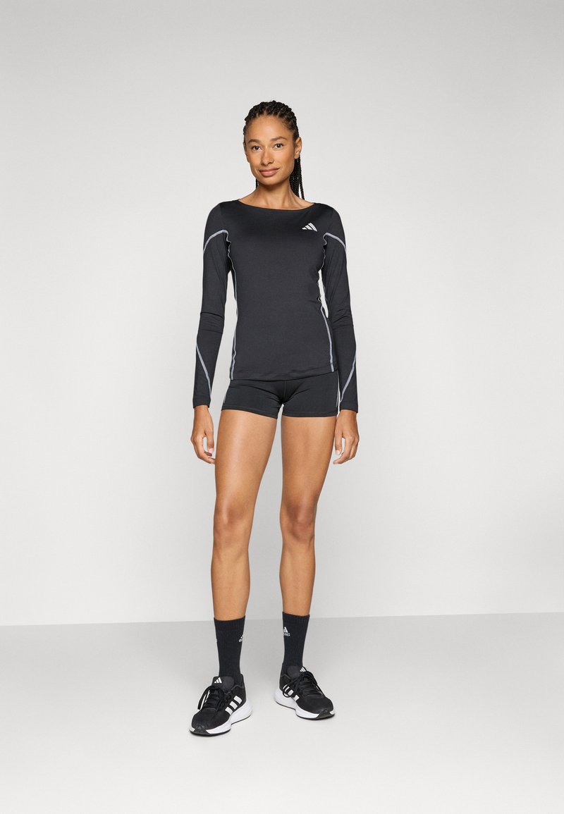 Schwarzes langärmliges Sportoberteil mit weißen Akzenten, kombiniert mit schwarzer Shorts und schwarzen Sportsocken. Das Modell trägt schwarze Laufschuhe.