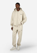 Mann in beigem Kapuzenzipper und passender Sweatpants mit Sneakers, der mit einer Hand in der Tasche vor einem weißen Hintergrund steht.