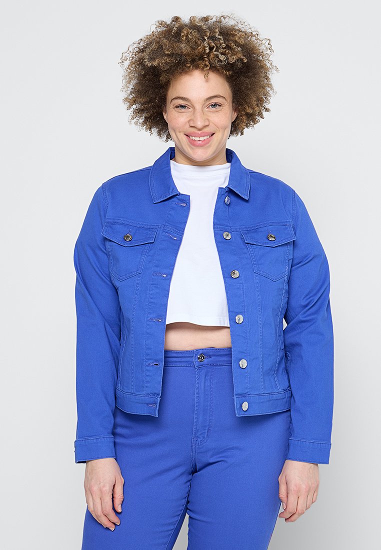 Blaue Jeansjacke mit klassischem Kragen, Fronttaschen und silbernen Knopfakzenten, kombiniert mit einem weißen Crop-Shirt und passenden blauen Hosen.