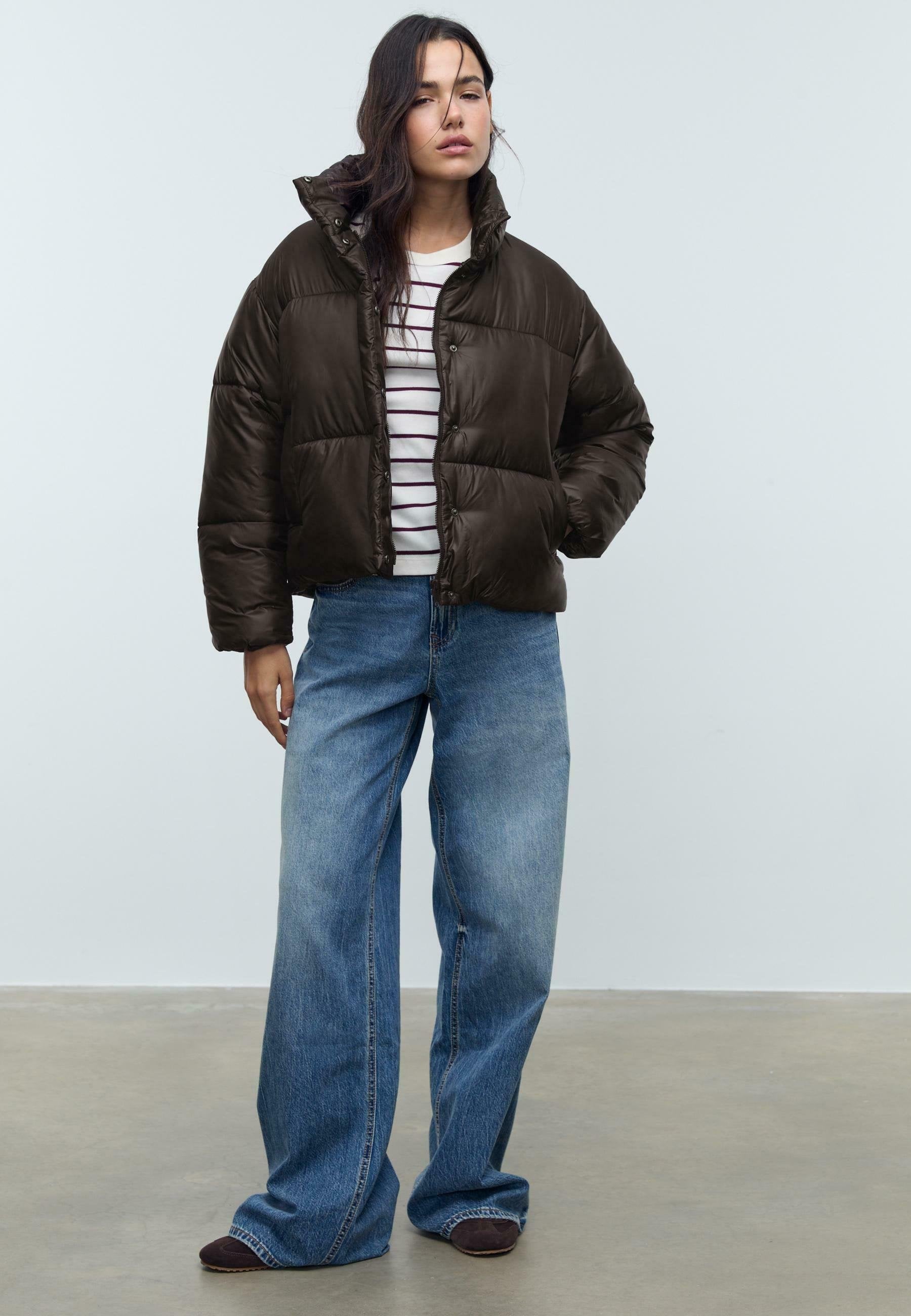 Frau in einer dunkelbraunen Steppjacke, gestreiftem weißen Shirt, weit geschnittenen blauen Jeans und dunklen Schuhen, die mit einer Hand in der Jackentasche steht.
