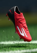 Roter und schwarzer Fußballschuh mit weißem Logo, in der Luft über einem Grasfeld mit aufgewirbeltem Rasen und sichtbarer weißer Begrenzungslinie.