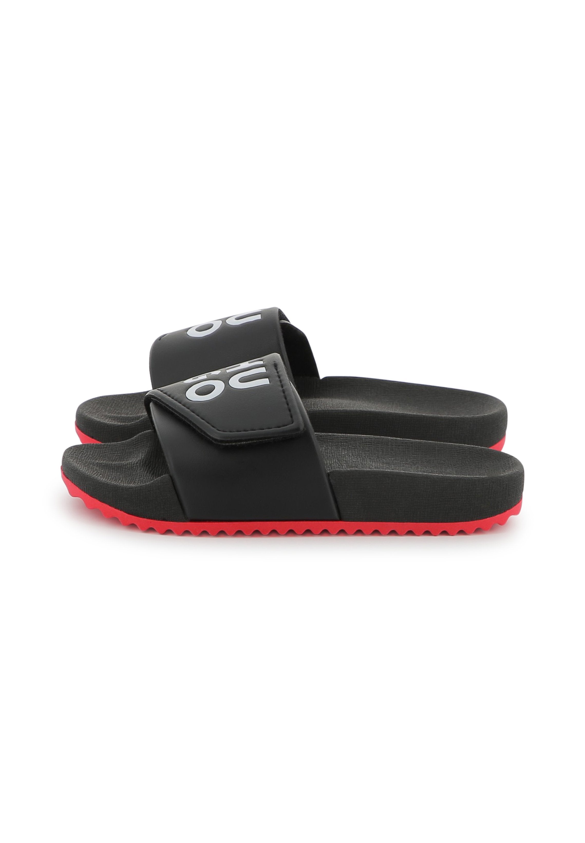 Schwarze Rutsch-Sandalen mit einem strukturierten Riemen, der ein auffälliges weißes Logo zeigt. Rote Gummisohle mit einem Zickzackmuster für besseren Halt.