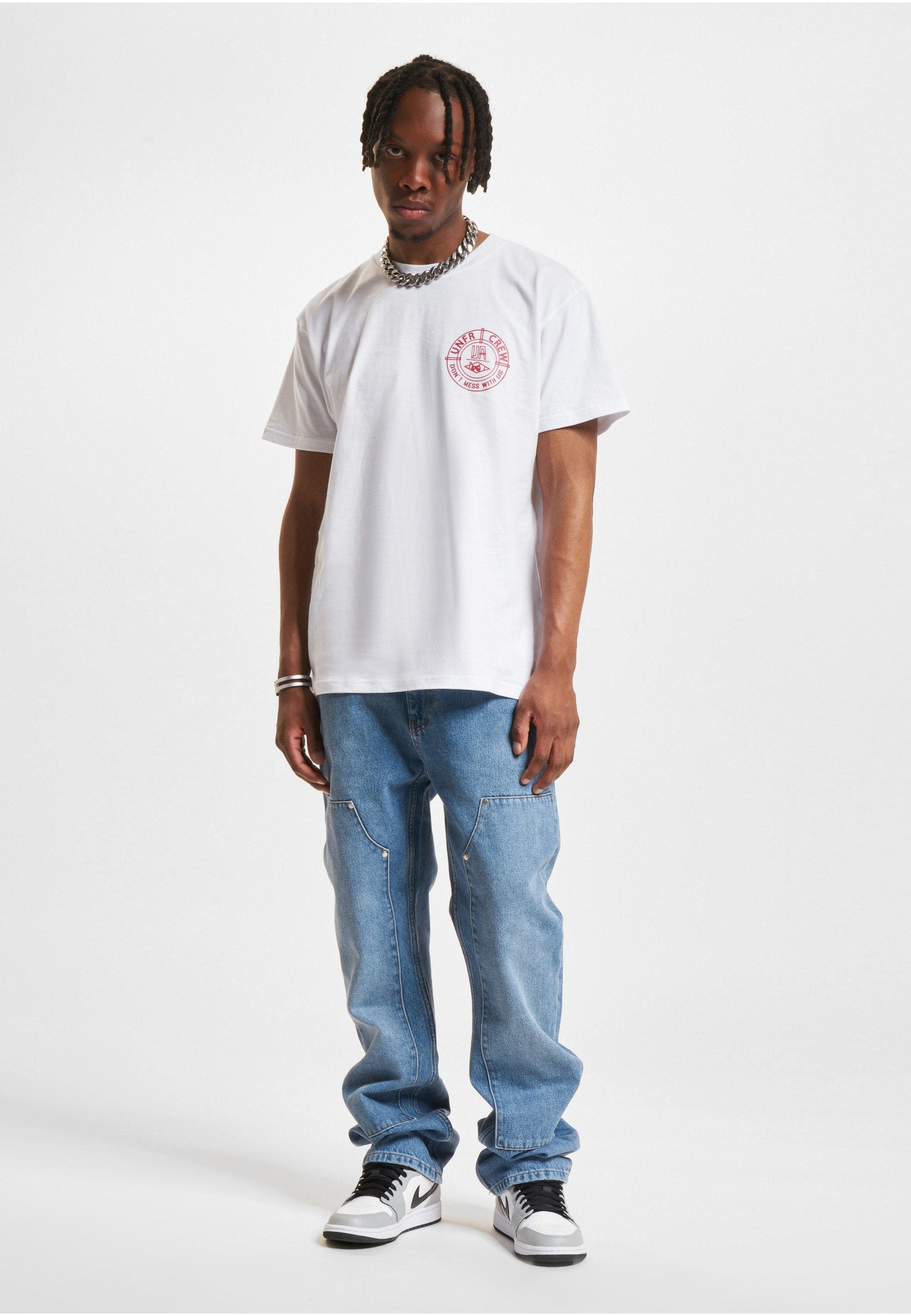 Mann mit geflochtenem Haar, der ein weißes T-Shirt mit Grafikmuster, weite hellblaue Jeans, weiße und schwarze Turnschuhe sowie eine silberne Kette trägt.