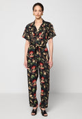 Schwarzer Jumpsuit mit kurzen Ärmeln und floralem Muster in Grün, Rot und Beige. Mit gebundenem Taillenbund und zwei Fronttaschen.