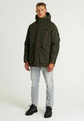 Olivengrüne gepolsterte Jacke mit Kapuze, zwei Fronttaschen und einer Reißverschlusstasche am Arm, kombiniert mit hellen Jeans und schwarzen Stiefeln.