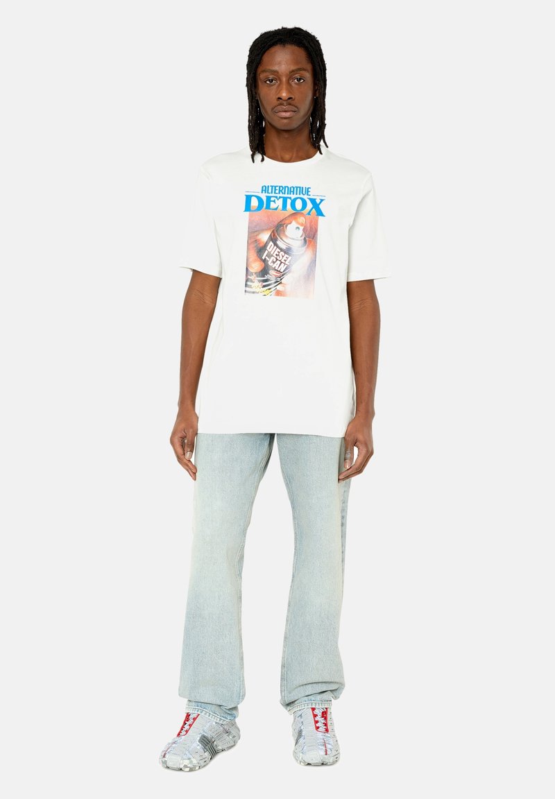 Weißes Baumwoll-T-Shirt mit einem bunten Grafikdruck, beschriftet mit "ALTERNATIVE DETOX" und einem Dosen-Design, kombiniert mit hellen Jeans und reflektierenden Schuhen.