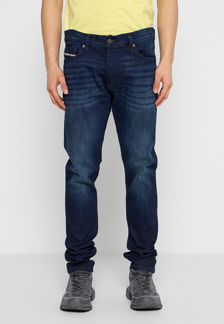 Dunkelblaue Jeans aus Denim mit schmaler Passform, Fronttaschen und strukturierter Oberfläche. Kombiniert mit grauen Sportschuhen.