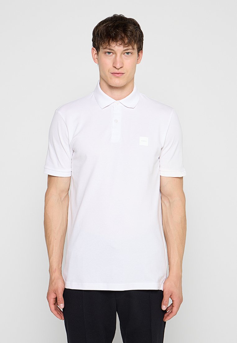Mann trägt ein schlichtes weißes Kurzarm-Poloshirt mit Kragen, steht vor einem einfarbigen, hellen Hintergrund, Hände entspannt an den Seiten.
