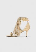 Goldene High Heel Sandalette mit Fransenverzierung, einem Knöchelriemen mit Schnalle und einem schlanken Stiletto-Absatz. Glänzende metallische Oberfläche.