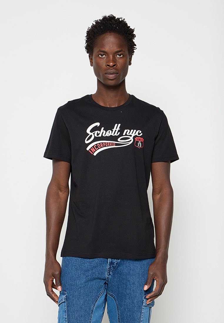 Junger Mann mit lockigem Haar trägt ein schwarzes "Schott nyc incorporated" T-Shirt und blaue Jeans-Cargohosen, steht vor einem schlichten hellen Hintergrund.