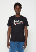 Junger Mann mit lockigem Haar trägt ein schwarzes "Schott nyc incorporated" T-Shirt und blaue Jeans-Cargohosen, steht vor einem schlichten hellen Hintergrund.
