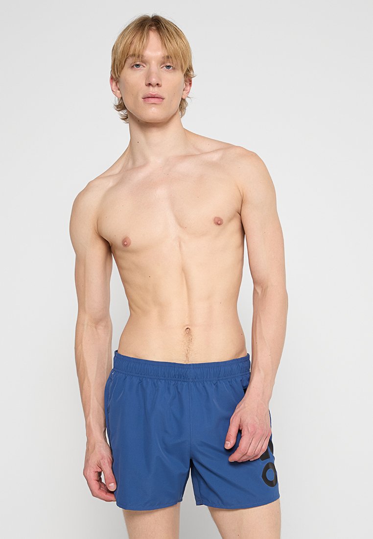 Blaue Badehose aus leichtem Stoff, mit einem elastischen Bund und einem großen schwarzen Logo am linken Bein. Minimalistisches Design.