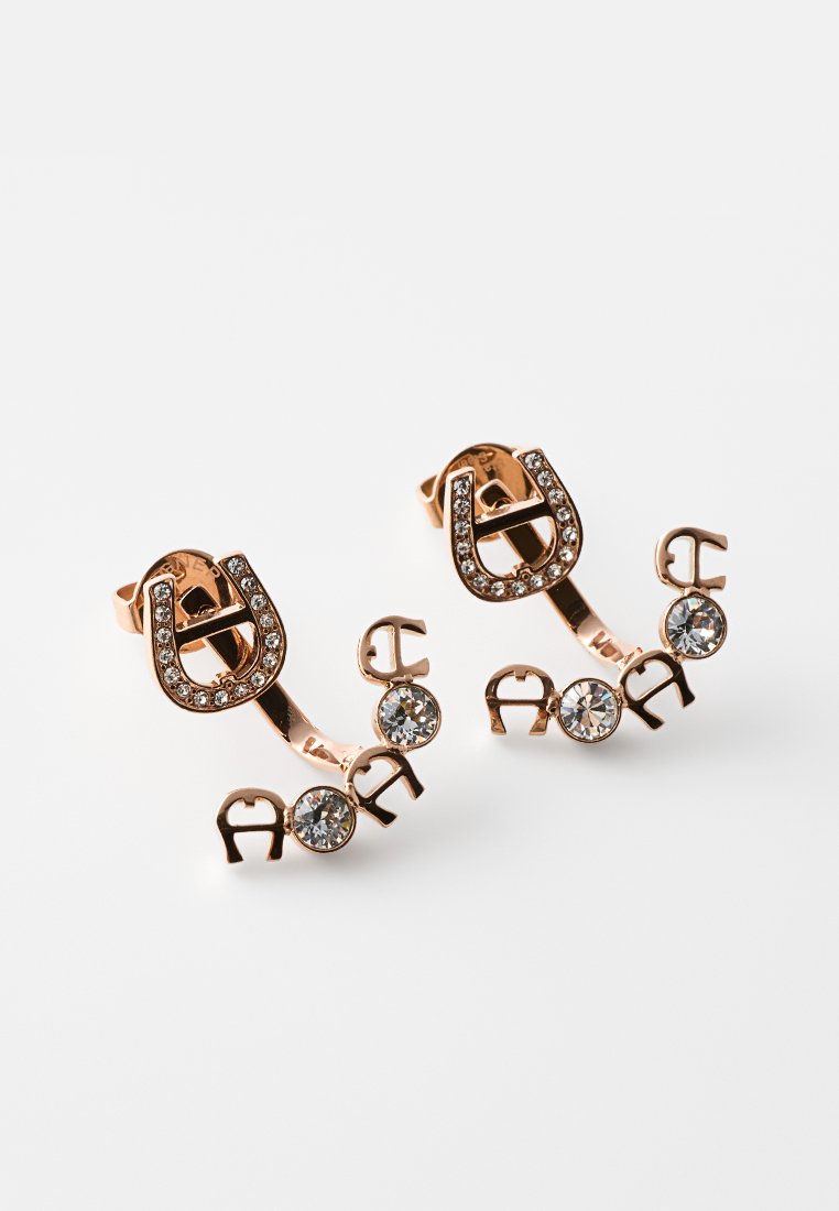 Roségold-Ohrringe mit Hufeisensteckern, besetzt mit kleinen Kristallen, und einer gebogenen Stange mit runden klaren Edelsteinen und Metallbuchstaben.