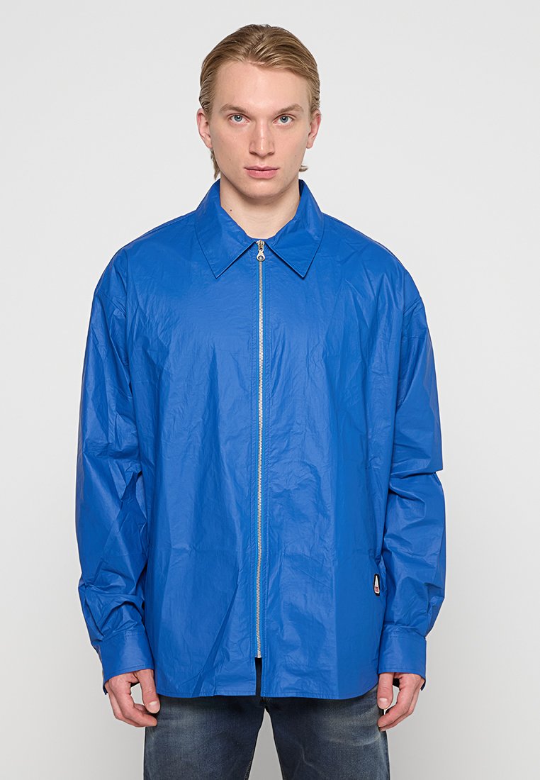 Leichte blaue Jacke mit Reißverschluss, Rückenschnitt, langen Ärmeln und einem dezenten Glanz. Mit kleinem Logo am Saum.