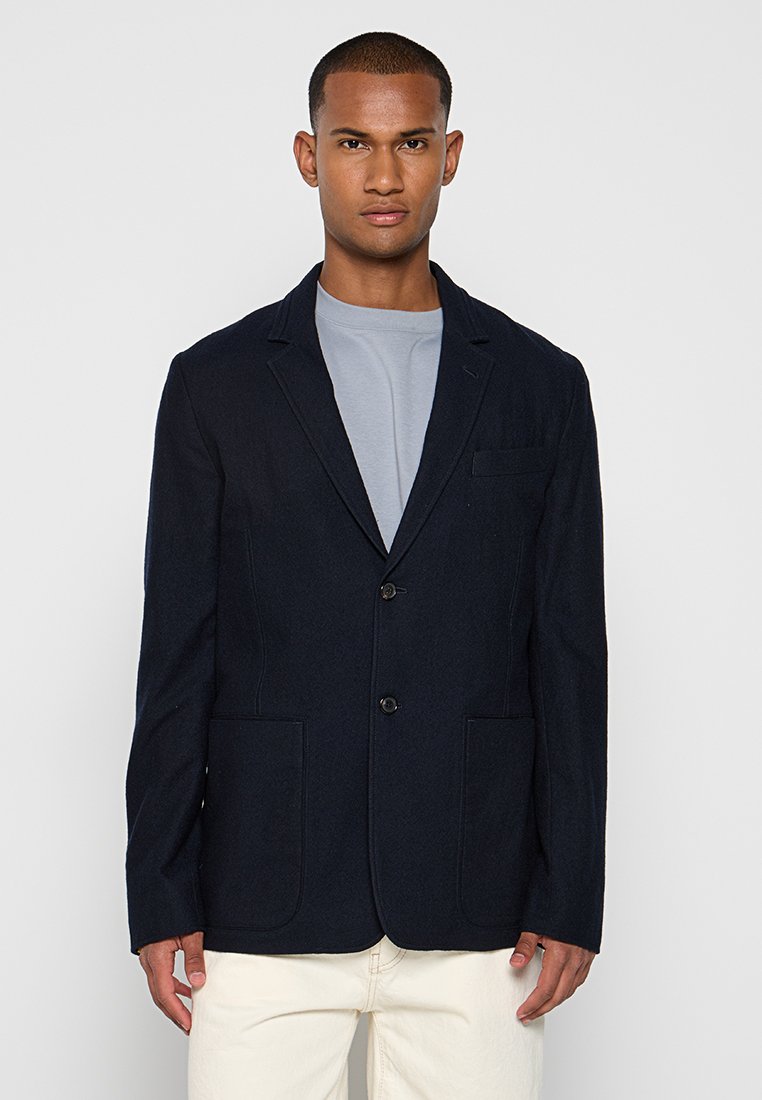 Ein Mann steht selbstbewusst da, trägt einen dunklen Blazer über einem hellgrauen T-Shirt, mit den Händen an seinen Seiten und einem neutralen Hintergrund.