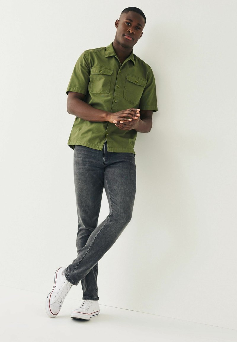 Olivegrühe Kurzarmhemd mit zwei Brusttaschen, kombiniert mit dunkelgrauen Slim-Fit-Jeans und weißen Sneakers.