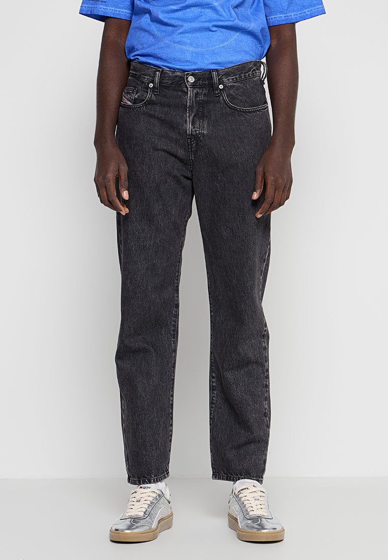 Person trägt schwarze Jeans mit geradem Bein, ein blaues Hemd und weiße Sneaker mit beigen Sohlen, steht vor einem schlichten hellen Hintergrund.