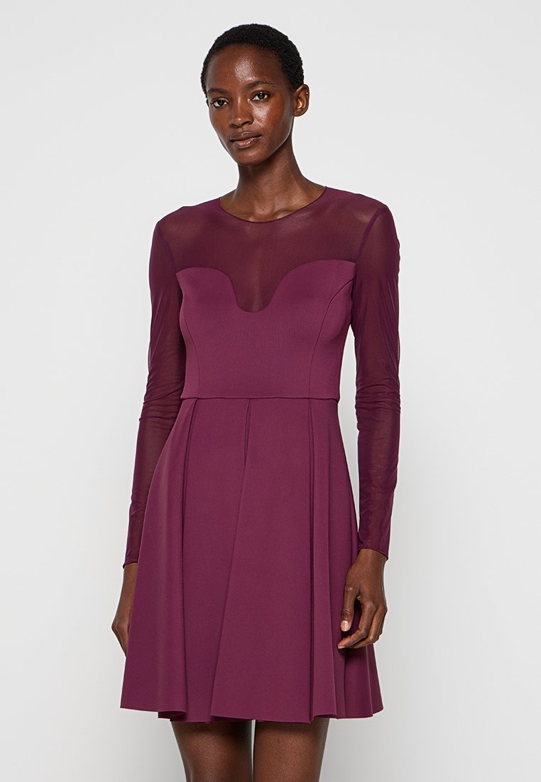 Frau in einem knielangen, pflaumenfarbenen Kleid mit transparenten langen Ärmeln und einem transparenten Oberteil, das vor einem schlichten Hintergrund steht.