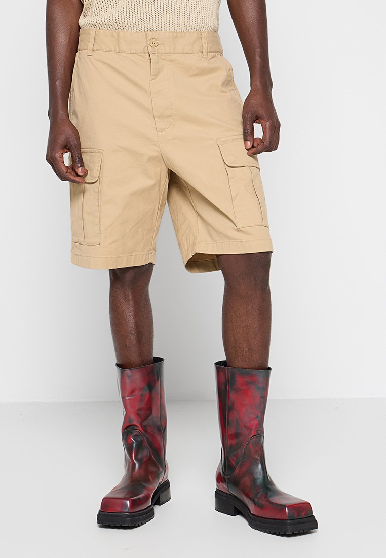 Khakifarbene Cargo-Shorts mit mehreren Taschen, Knopfverschluss und entspanntem Schnitt, kombiniert mit knielangen, schwarz-rot marmorierten Gummistiefeln.