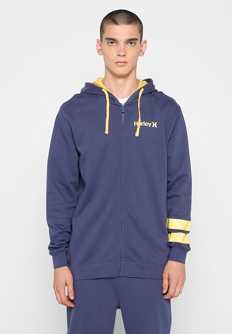 Junger Mann mit kurzen Haaren, der einen dunkelblauen Reißverschluss-Hoodie mit gelben Kordeln und gelben Streifen auf dem linken Ärmel trägt, steht vor einem weißen Hintergrund.