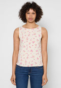Frau mit lockigem Haar, die ein ärmelloses cremefarbenes Oberteil mit kleinem Muster aus rosa und blauen Blumen und eine blaue Jeans trägt, stehend vor einem einfarbigen Hintergrund.