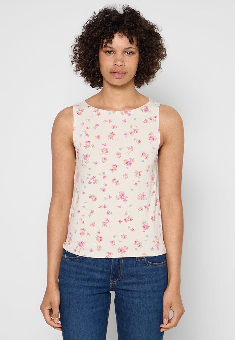 Frau mit lockigem Haar, die ein ärmelloses cremefarbenes Oberteil mit kleinem Muster aus rosa und blauen Blumen und eine blaue Jeans trägt, stehend vor einem einfarbigen Hintergrund.