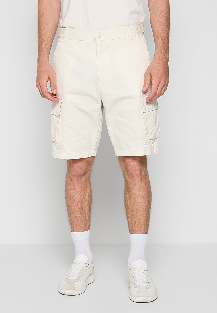 Mann in beigen Cargo-Shorts, weißen Kniestrümpfen und weißen Turnschuhen, der vor einem schlichten Hintergrund steht.