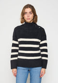 Frau mit schulterlangem Haar, die einen schwarzen Strickpullover mit weißen horizontalen Streifen und Knopfdetails an der Schulter trägt, kombiniert mit blauen Jeans.