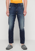Dunkelblaue Jeans mit schmaler Passform, leichter Ausbleichung an den Knien und elastischem Bund mit Kordelzug. Getragen mit grauen Slippern.