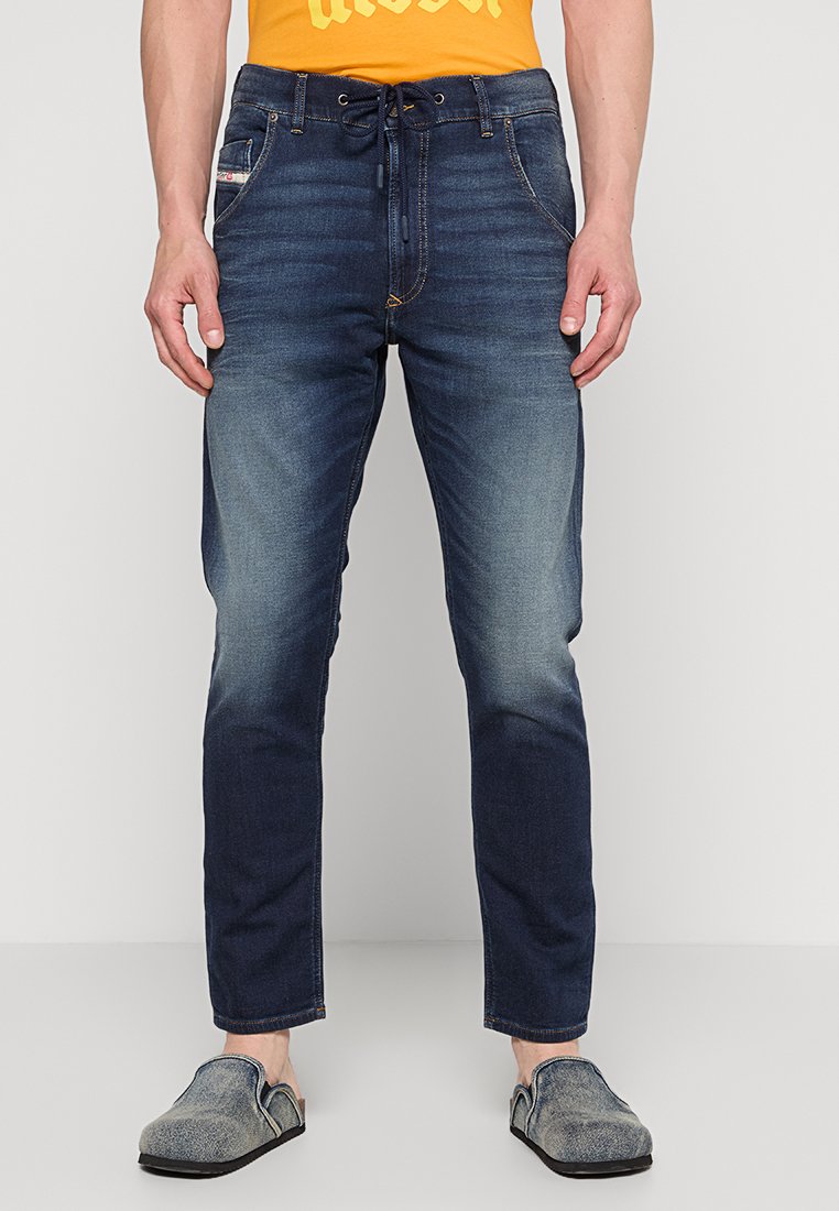 Dunkelblaue Jeans mit schmaler Passform, leichter Ausbleichung an den Knien und elastischem Bund mit Kordelzug. Getragen mit grauen Slippern.