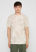 Junger Mann mit kurzem dunklem Haar, trägt ein beige gemustertes kurzärmliges Hemd und olivgrüne Hose, steht vor einem einfarbigen hellen Hintergrund.