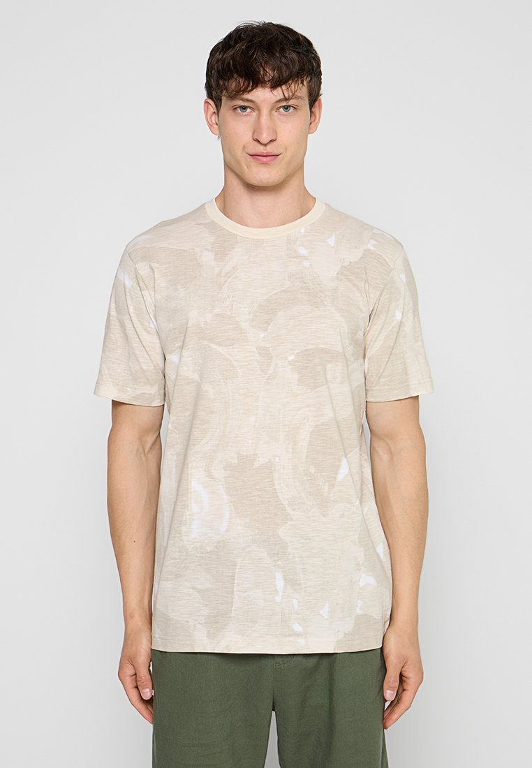 Junger Mann mit kurzem dunklem Haar, trägt ein beige gemustertes kurzärmliges Hemd und olivgrüne Hose, steht vor einem einfarbigen hellen Hintergrund.
