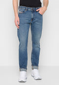 Mann in mid-blauen aufgerollten Jeans, schwarzem Hemd und weißen Sneakern, der mit einer Hand in der Tasche vor einem schlichten Hintergrund steht.