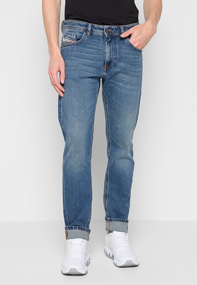 Mann in mid-blauen aufgerollten Jeans, schwarzem Hemd und weißen Sneakern, der mit einer Hand in der Tasche vor einem schlichten Hintergrund steht.