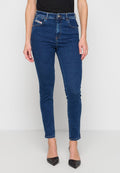 Frau trägt dunkelblaue Skinny-Jeans, schwarze spitze Absätze und ein schwarzes Oberteil, steht vor einer weißen Wand und einem weißen Boden.
