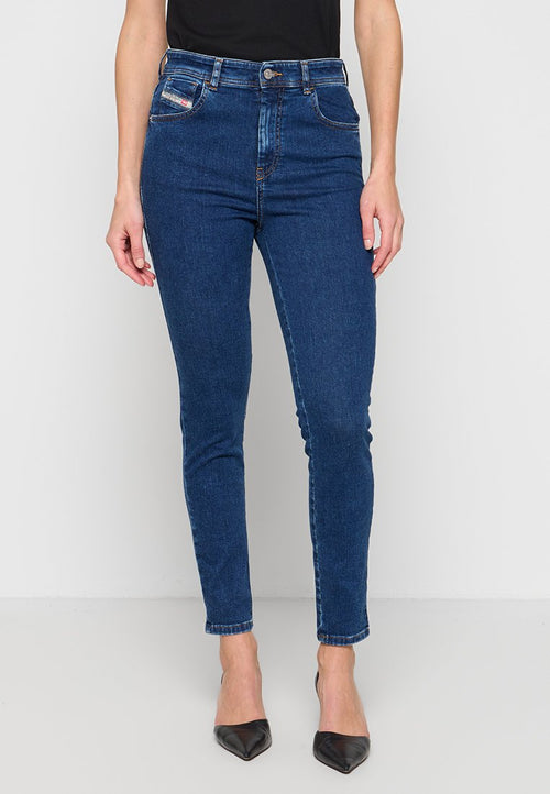 Frau trägt dunkelblaue Skinny-Jeans, schwarze spitze Absätze und ein schwarzes Oberteil, steht vor einer weißen Wand und einem weißen Boden.