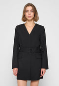 Frau mit kurzen, hellbraunen Haaren, die ein schwarzes, langärmeliges Blazer-Kleid mit Gürtel trägt, stehend vor einem schlichten, hellen Hintergrund.