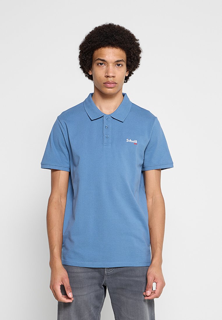 Junger Mann mit lockigem Haar, der ein blaues Polohemd mit kleinem weiß-rotem Logo auf der Brust und graue Jeans trägt, vor einem einfarbigen Hintergrund steht.