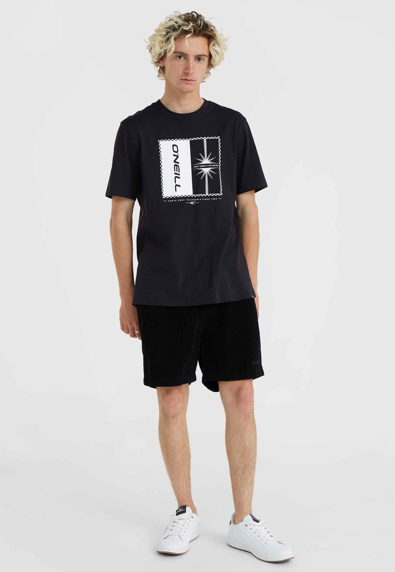Schwarzes Baumwoll-T-Shirt mit grafischem Druck im Sternen-Motiv, kombiniert mit schwarzen Cordshorts und weißen Sneakers.