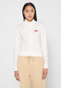 Weiße Zip-Jacke mit hohem Kragen, ausgestattet mit einem kleinen roten Diamant-Logo auf der linken Seite. Weicher Stoff, taillierte Passform, kombiniert mit beigen Hosen.