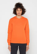 Frau mit lockigem Haar, die ein knalloranges Sweatshirt mit langen Ärmeln und schwarze Hose trägt, vor einem schlichten weißen Hintergrund steht.