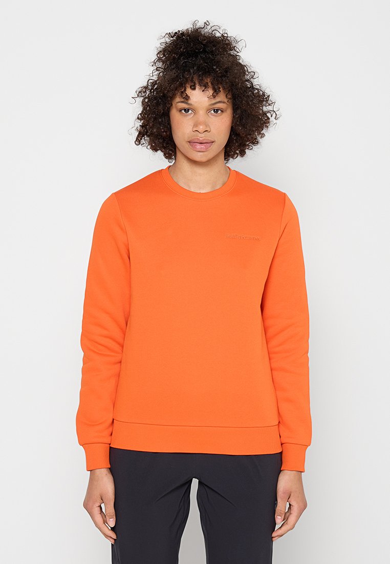 Frau mit lockigem Haar, die ein knalloranges Sweatshirt mit langen Ärmeln und schwarze Hose trägt, vor einem schlichten weißen Hintergrund steht.
