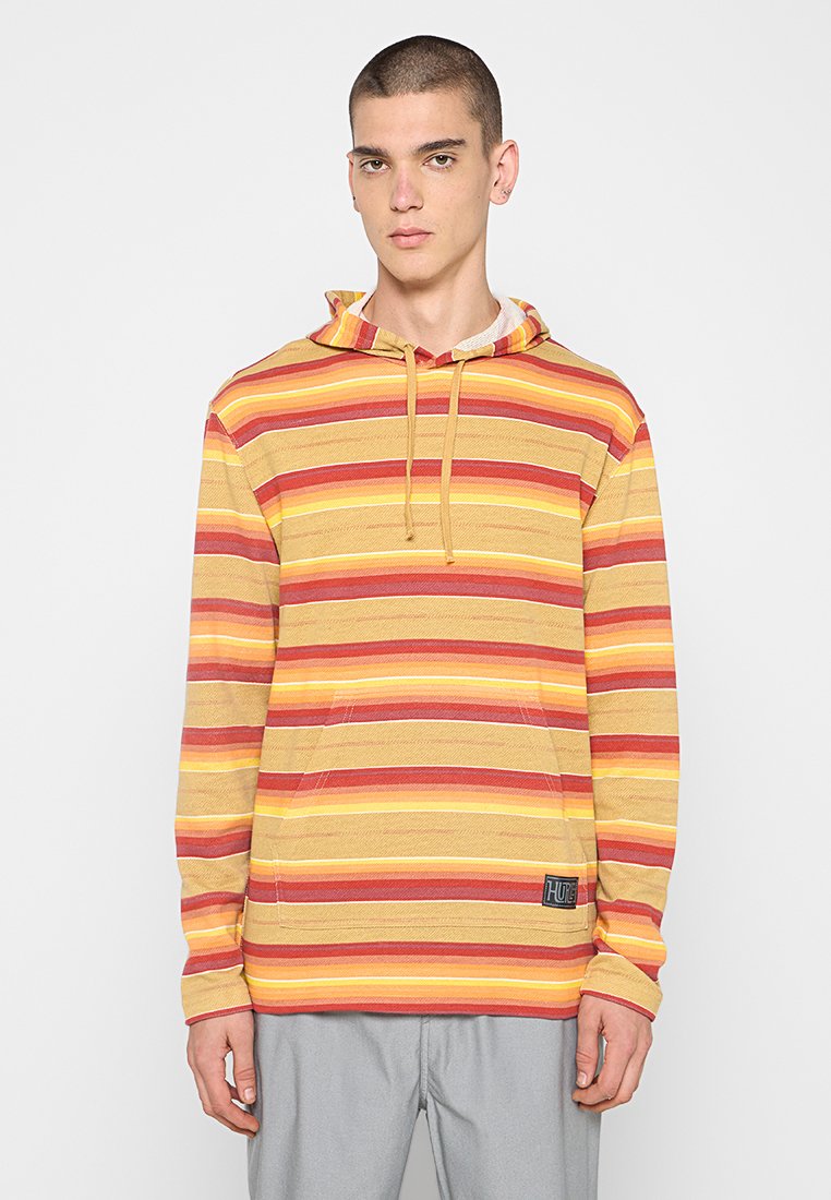 Junger Mann in einem gelb, rot und orange gestreiften Kapuzenpullover mit Fronttasche und hellgrauen Hosen, stehend vor einem weißen Hintergrund.