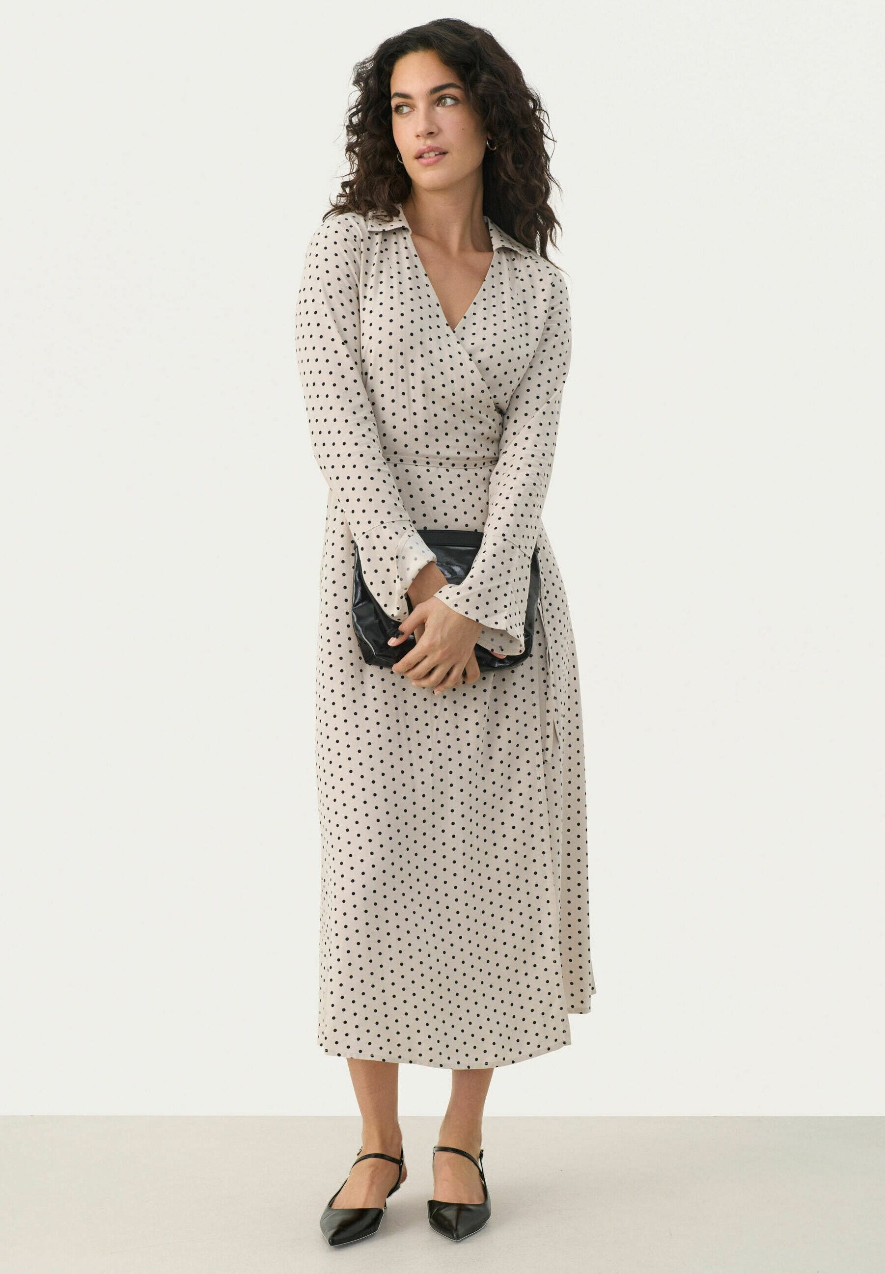 Beige Wickelkleid mit weißen Punkten, langen Ärmeln und V-Ausschnitt, kombiniert mit schwarzen spitzen Ballerinas und einer schwarzen Clutch.