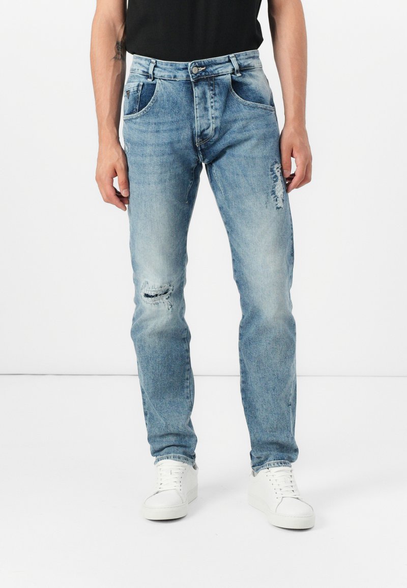 Hellblaue Designerjeans mit Distressed-Details, fünf Taschen, gerader Schnitt, kombiniert mit weißen Sneakern und einem schwarzen Shirt.