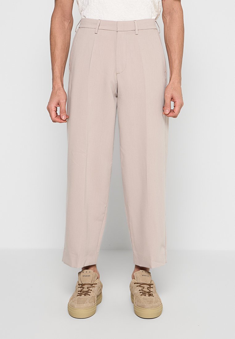 Mann trägt beige, gekürzte, maßgeschneiderte Hosen und passende beige Sneaker und steht vor einem schlichten Hintergrund.