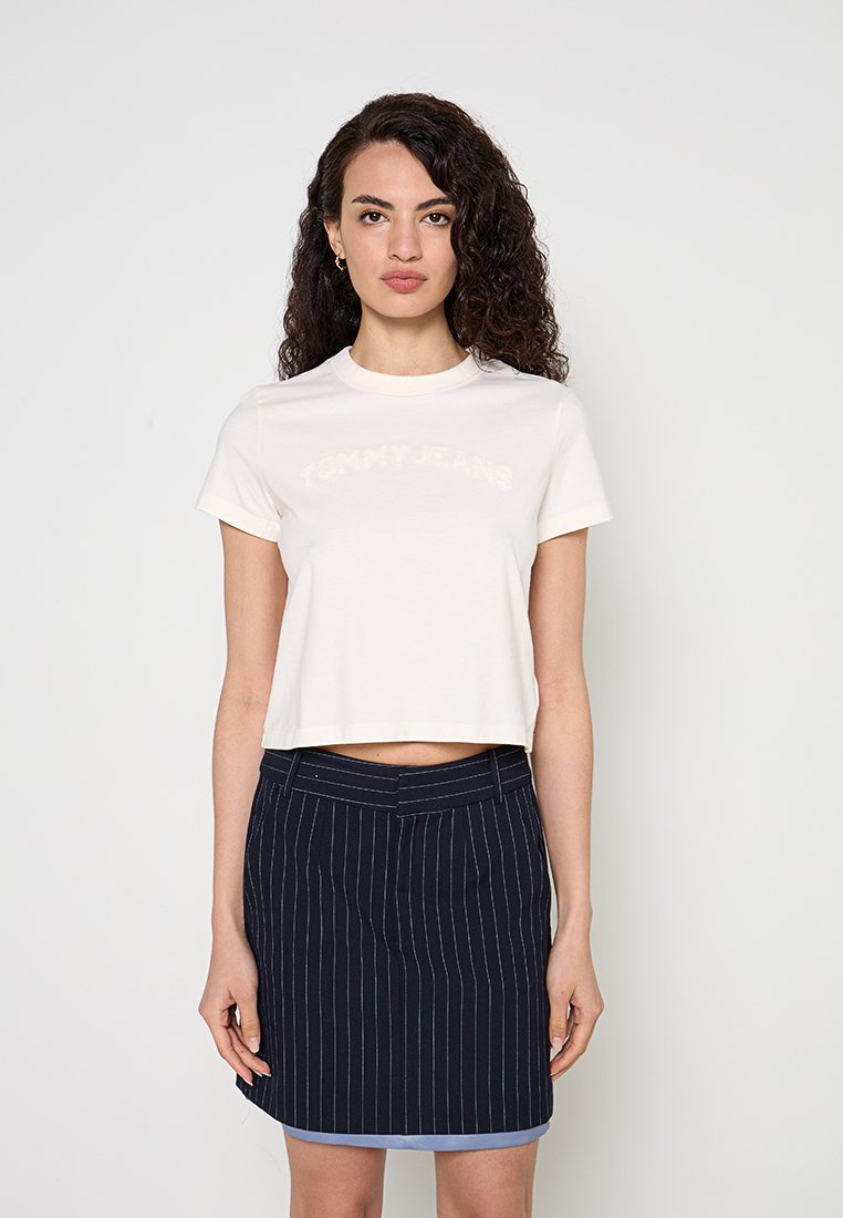 Frau mit lockigem Haar, die ein weißes bauchfreies T-Shirt und einen dunkelblauen Rock mit Nadelstreifen vor einem schlichten hellen Hintergrund trägt.